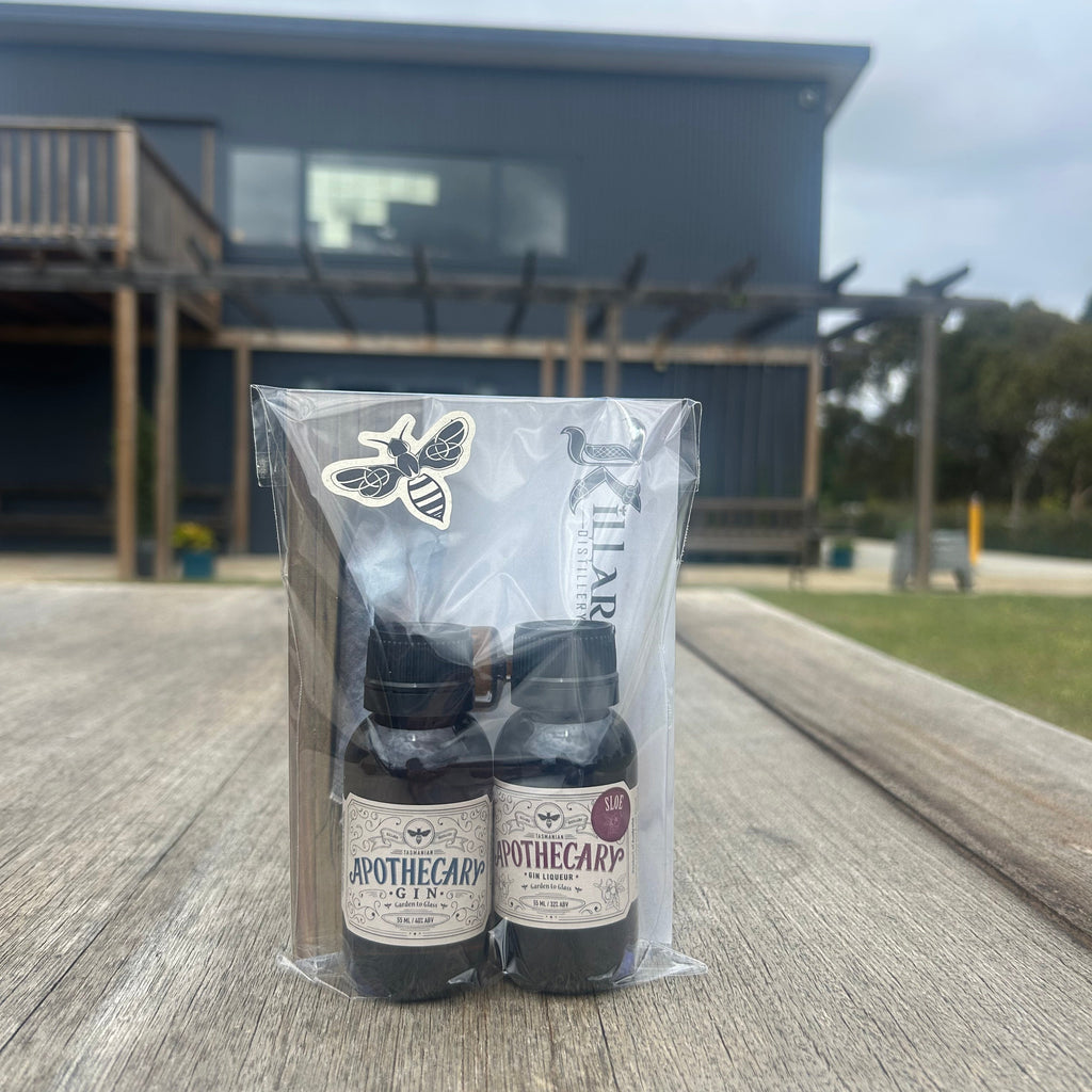 Two small bottles of Apothecary Gin Range in a clear plastic bag on a wooden surface with a building in the background.